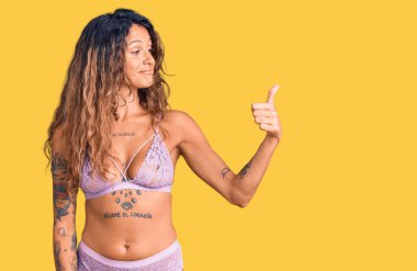 Dövmeli, iç çamaşırlı, gururlu, gülümseyen, baş parmak kaldıran genç İspanyol kadın. 