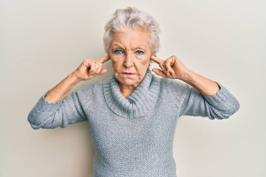 Yaşlı, kır saçlı, kulaklarını parmaklarıyla kapatan, şüpheci ve gergin bir kadın sorun yüzünden kaşlarını çatıyor. negatif kişi. 