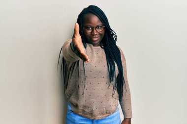 Saçları örgülü, günlük kıyafetler giyen ve gözlüklü genç siyahi bir kadın dostça gülümseyerek selamlama ve hoş geldin diyor. başarılı iş. 