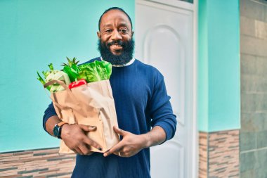 Sakallı Afrikalı Amerikalı bir adam elinde süpermarketten alınmış kağıt poşetler tutuyor.
