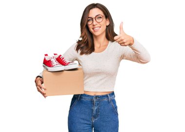 Genç esmer kadın kırmızı ayakkabılı kutu tutuyor mutlu ve pozitif gülümsüyor, başparmağıyla mükemmel ve onay işareti yapıyor. 
