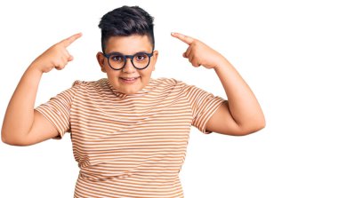 Gündelik kıyafetler ve gözlük takan küçük çocuk iki eliyle başını gösteren gülümseme, harika fikir ya da düşünce, iyi hafıza 