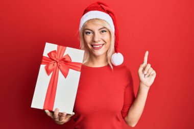 Genç sarışın kız Noel şapkası takıyor ve elinde bir hediyeyle bir fikir veya soruyla gülümsüyor. 