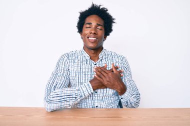 Yakışıklı Afro saçlı, sıradan kıyafetler giyen, göğsünde elleri kapalı, yüzünde minnet dolu bir jestle masada oturan Afro-Amerikan erkeği. sağlık kavramı. 