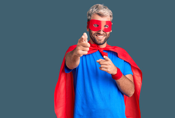 Young blond man wearing super hero custome pointing fingers to camera with happy and funny face. good energy and vibes. 