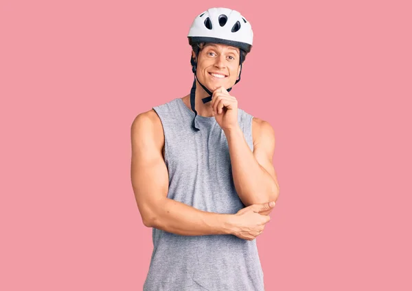 Young handsome man wearing bike helmet looking confident at the camera with smile with crossed arms and hand raised on chin. thinking positive.