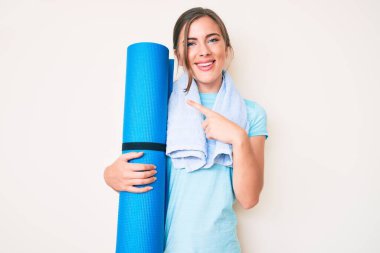 Güzel, beyaz bir kadın yoga minderi tutuyor, gülümsüyor, mutlu bir şekilde eli ve parmağı işaret ediyor. 