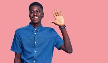 Genç, Afro-Amerikan bir adam, günlük giysiler içinde mutlu ve gülümseyen bir merhaba diyor. 