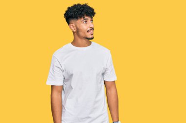 Gündelik beyaz tişört giyen genç bir Arap yüzünde gülümseme, doğal bir ifade ile yan yana bakıyor. Kendinden emin gülen. 