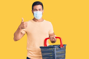 Alışveriş sepeti ve tıbbi maske takan genç İspanyol adam mutlu ve olumlu gülümsüyor, başparmağı mükemmel ve onay işareti yapıyor. 