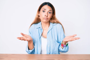 Masada oturan esmer, sıradan kıyafetler giyen, kafası karışmış, elleri ve kolları havada genç bir kadın. şüphe kavramı. 