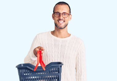 Süpermarket alışveriş sepeti tutan yakışıklı genç adam pozitif ve mutlu görünüyor. Duruyor ve gülümsüyor. Kendine güvenen bir gülümsemeyle dişlerini gösteriyor. 