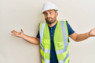 Güvenlik kaskı takan sakallı, parlak ceketli, kolları açık ve kafası karışık yakışıklı bir adam.. 