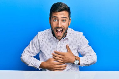 İş kıyafetleri giymiş yakışıklı İspanyol adam masaya oturmuş gülümsüyor ve kahkahalarla gülüyor çünkü elleri vücutta komik bir şaka var.. 