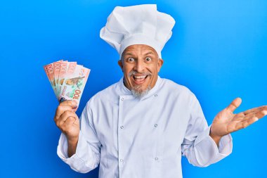 Orta yaşlı, gri saçlı, profesyonel aşçı üniforması giyen elinde yeni Zelanda doları banknotları olan mutlu bir gülümsemeyle başarıyı kutlayan ve kazanan yüz ifadesiyle elini kaldıran bir adam. 