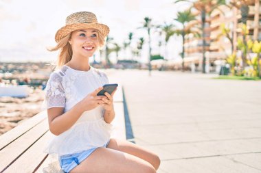 Kafkasyalı genç turist mutlu bir şekilde gülümsüyor akıllı telefon kullanıyor gezinti güvertesinde bankta oturuyor..