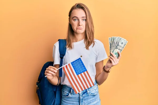 Beautiful blonde woman exchange student holding usa flag and dollars banknotes depressed and worry for distress, crying angry and afraid. sad expression.