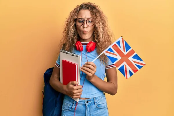 Beautiful caucasian teenager girl exchange student holding uk flag making fish face with mouth and squinting eyes, crazy and comical.