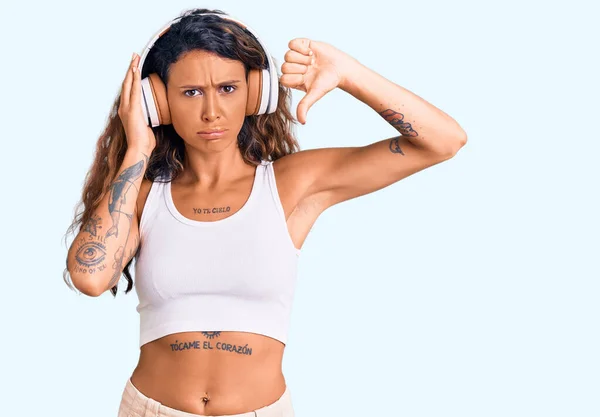 Young hispanic woman with tattoo listening to music using headphones with angry face, negative sign showing dislike with thumbs down, rejection concept