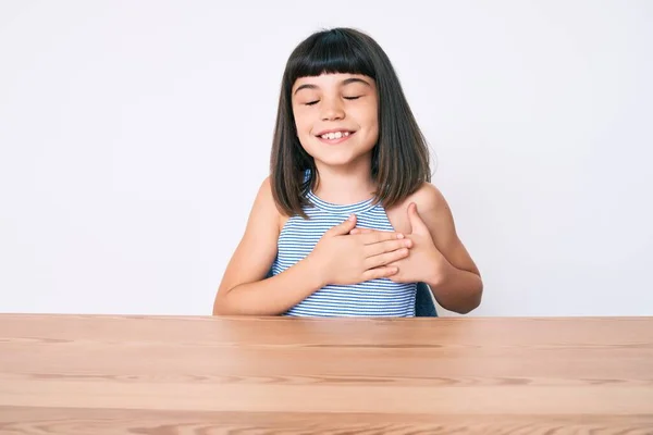 年轻的小女孩穿着休闲装 手放在胸前笑着坐在桌子上 满脸感激地闭上眼睛 健康概念 — 图库照片
