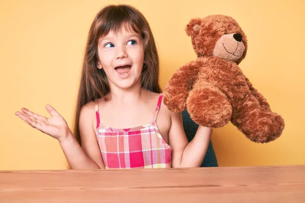 一个有着长发的高加索小女孩坐在桌子上 泰迪熊带着快乐的微笑和高傲的表情庆祝胜利 — 图库照片