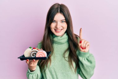 Genç, güzel, beyaz bir kız elinde pasta tatlısı tutarken gülümsüyor bir fikirle ya da soru işareti yaparak mutlu bir yüzle, bir numara 