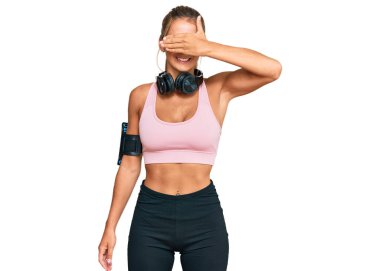 Spor kıyafetleri giyen, kulaklık takan, gülümseyen ve gülümseyen genç sarışın kadın şaşırtmak için gözlerini kapatıyor. kör kavram. 