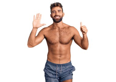 İspanyol asıllı genç adam mayo giyiyor. Altıncı parmaklarıyla gösteriş yapıyor ve gülümsüyor. Kendinden emin ve mutlu.. 