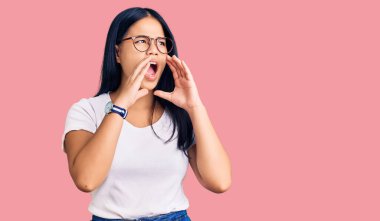 Genç, güzel Asyalı bir kız günlük kıyafetler ve gözlük takıyor ve elleri ağzında bağırıyor. 