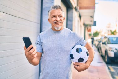 Orta yaşlı, ispanyol, kır saçlı, akıllı telefon kullanan ve şehirde futbol topu tutan bir adam..