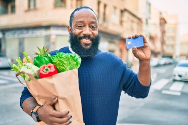 Sakallı Afrikalı Amerikalı bir adam elinde süpermarketten alınmış kağıt torbalarla kredi kartı gösteriyor.