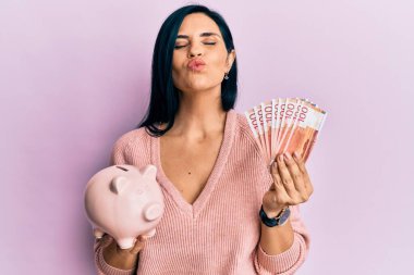 Genç beyaz kadın Norveçli krona banknotları tutuyor ve kumbarası kameraya bakıyor. Öpücük üflüyor, sevimli ve seksi. aşk ifadesi. 