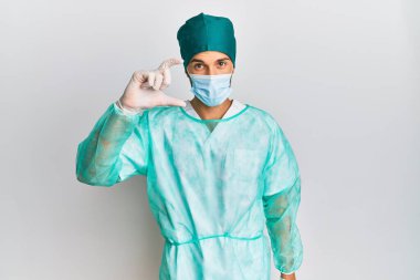 Genç, yakışıklı, cerrah üniforması giyen, tıbbi maske takan, gülümseyen, kendine güvenen, el hareketi yapan, parmaklarıyla işaret eden ve kameraya bakan bir adam. ölçüm kavramı. 
