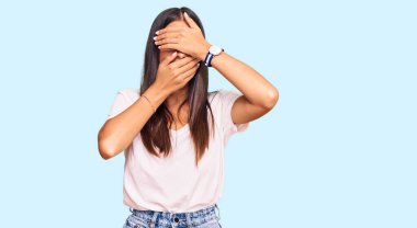 Günlük beyaz tişört giyen genç İspanyol kadın gözlerini ve ağzını elleriyle kapatıyor, şaşırmış ve şok olmuş. duygu gizleme 