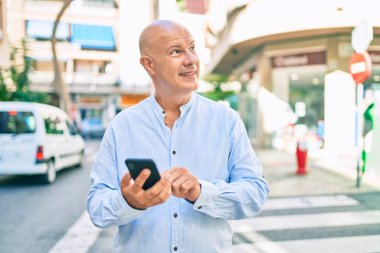Orta yaşlı kel adam şehirde akıllı telefon kullanarak mutlu bir şekilde gülümsüyor..