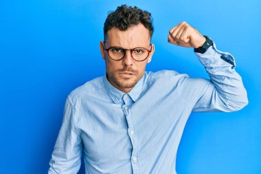 Sakallı, sıradan giyinmiş ve gözlüklü yakışıklı. Kollarını kavuşturup arkasını kolluyor. 