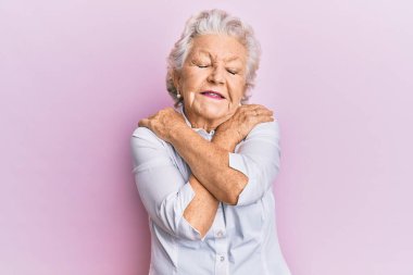 Kıdemli, gri saçlı, günlük kıyafetler giyen, kendine sarılan mutlu ve pozitif, kendinden emin gülümseyen bir kadın. Sevgi ve özgüven. 