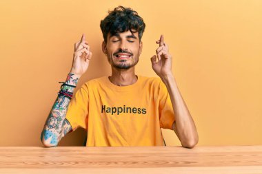 Üzerinde mutluluk mesajı olan tişört giyen genç İspanyol adam masaya oturmuş umut ve gözlerin kapalı bir şekilde gülümseyerek el sallıyor. Şans ve batıl inançlar. 