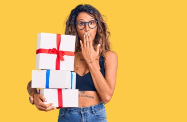 Dövmeli genç bir İspanyol kadın. Elinde hediyeler var. Ağzını eliyle kapatıyor. şaşırmış ifade 