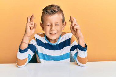 Gündelik giysiler giymiş sevimli beyaz çocuk masanın üzerinde oturmuş umut ve gözler kapalı bir şekilde gülümseyerek el sallıyor. Şans ve batıl inançlar. 