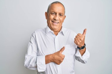 Yakışıklı, beyaz tişörtlü, arka tarafı işaret eden, el ve baş parmakları havada, kendinden emin bir şekilde gülümseyen bir adam. 