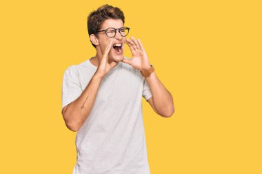 Yakışıklı beyaz adam, günlük kıyafetler ve gözlük takıyor ve elleri ağzında bağırıyor. 