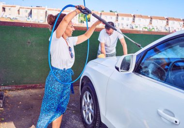 Orta yaşlı, güzel bir çift günlük kıyafetler giyip mutlu gülümsüyor. Yüzünde gülümseme, su tabancası ve sünger kullanarak araba yıkamak..