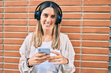 Beyaz kadın şehirde akıllı telefon ve kulaklık kullanarak mutlu bir şekilde gülümsüyor..