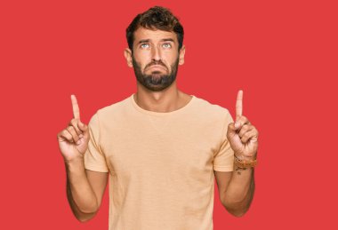 Sakallı, sıradan tişörtlü yakışıklı adam üzgün ve üzgün görünüyor, parmaklarıyla yönü gösteriyor, mutsuz ve depresif.. 