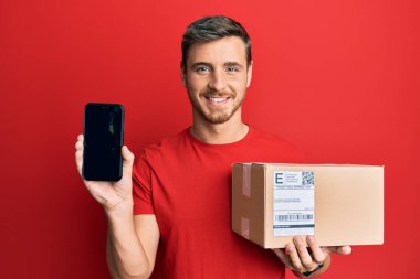 Yakışıklı beyaz adam teslimat paketini tutuyor ve yüzünde mutlu ve havalı bir gülümsemeyle akıllı telefon ekranı gösteriyor. diş gösterme. 