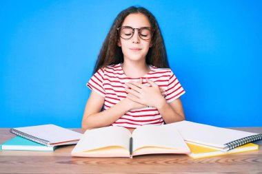 Sevimli İspanyol kız okul sınavına hazırlanıyor. Masada oturmuş gülümsüyor, elleri göğsünde, gözleri kapalı ve yüzünde minnet dolu bir jest var. sağlık kavramı. 