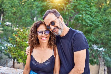 Orta yaşlı bir çift, taş duvarda oturup parktaki kameraya bakarken mutlu bir şekilde gülümsüyor.