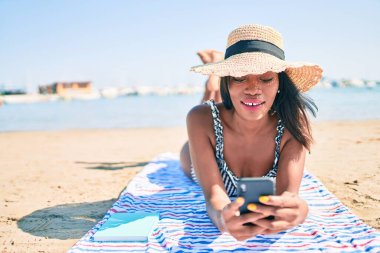 Genç Afro-Amerikan kadın tatilde. Havluya uzanmış, kumsalda akıllı telefon kullanıyor.