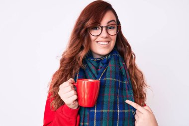 Kış eşarbı takan ve bir fincan sıcak kahve içen genç ve güzel bir kadın mutlu bir şekilde eli ve parmağı ile işaret ediyor. 